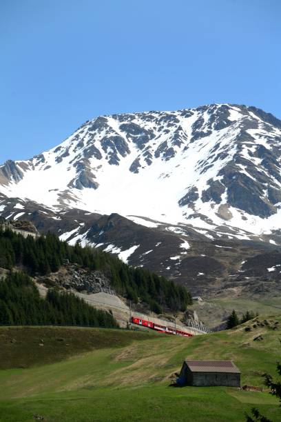 istock View of a red train passing through a green valley with a snowy mountain backdrop under a blue sky. 2192243401