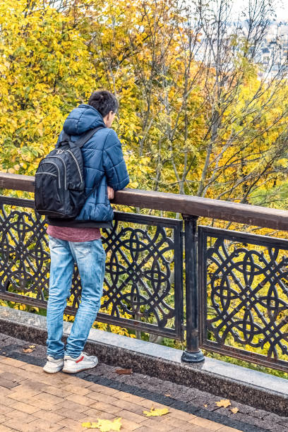 istock A man in a blue jacket and jeans stands at a railing, overlooking a lush park filled with vibrant autumn foliage in Kyiv, Ukraine 2192246834