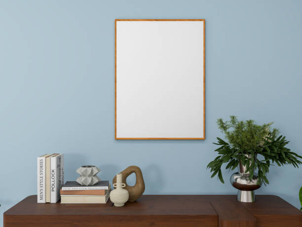 istock A minimalist interior scene featuring a blank wooden-framed picture frame on a white wall, a wooden table with books, decorative vases, and a metallic vase with green foliage. 3d rendering. 2192260677