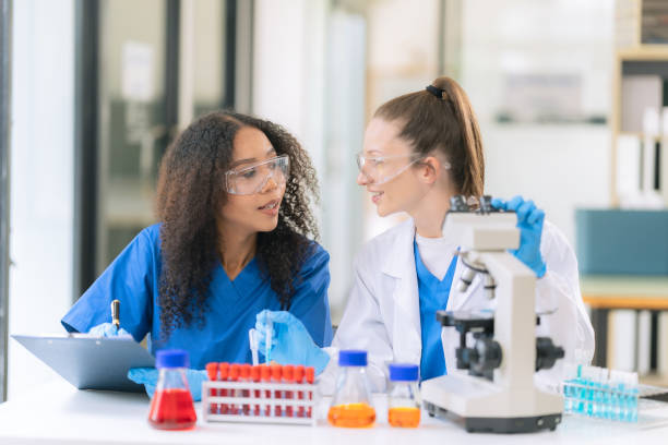 istock Two beautiful female doctors sitting at a table in a science lab with test flasks. Colorful glass bottles filled with various liquids on table with microscope 2192268392