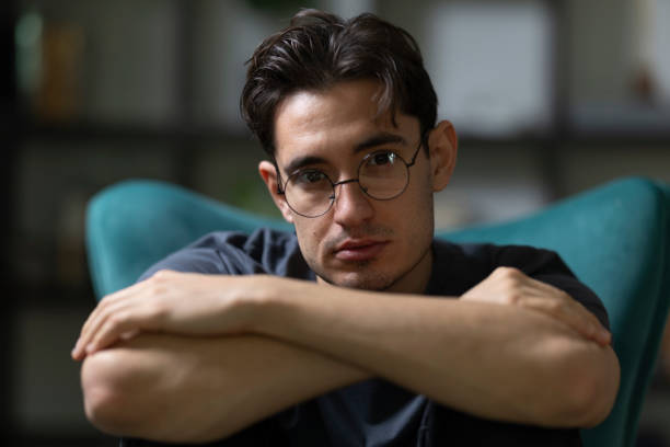 Portrait of a thoughtful young man with eyeglasses sitting on a modern chair with his arms crossed, conveying a sense of contemplation and introspection
