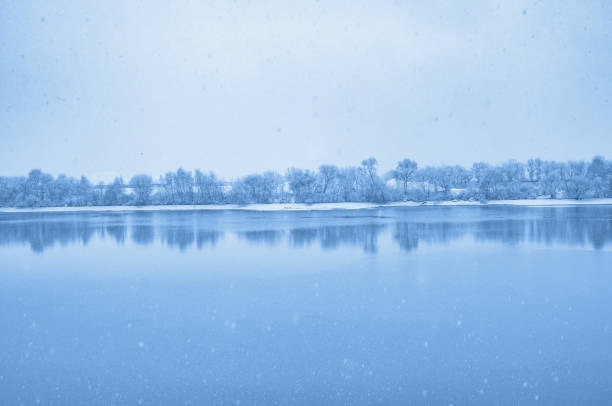 A calm river flows through a snowy winter landscape, with trees, reeds, and bushes covered in fresh snow as snowflakes gently fall.