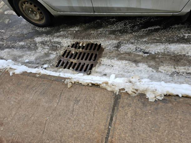 A drain with some snow and ice around it along the side of a sidewalk.