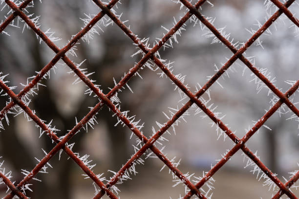 A close-up of a red metal fence covered in frost crystals, forming sharp, icy patterns. The blurred background enhances the geometric design and contrasts with the frosty details.