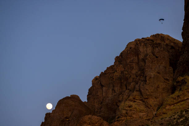 Paraglider soars over desert landscape at dusk, full moon rises behind cliff in distance