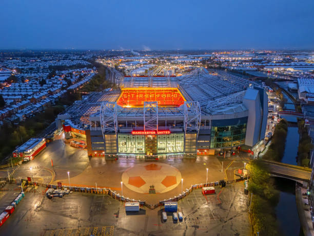 Manchester, UK - September 30, 2025: Modern stadium exterior in Manchester with red panels, grey pillars, glass entrance, dramatic sky.
