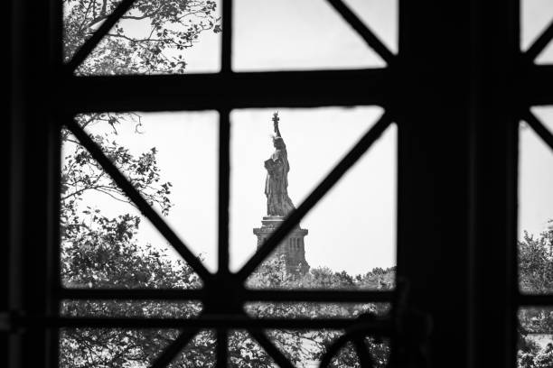 Black  and white photo of the Statue of Liberty taken at a distance, through a window at the Ellis Island National Museum of Immigration.