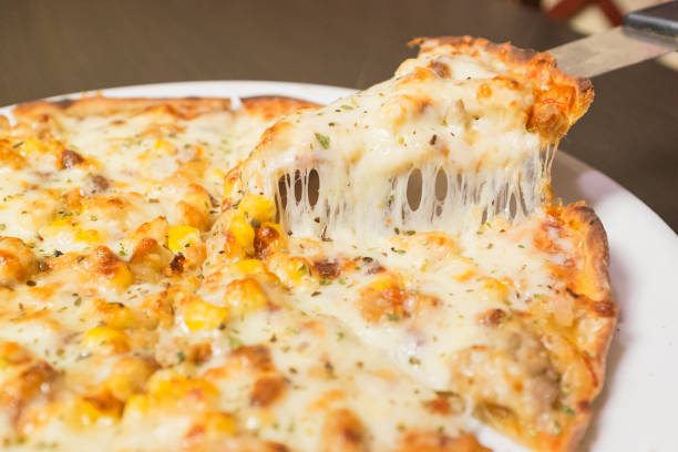 A slice of pizza with cheese and corn on it. A knife is being used to cut the pizza