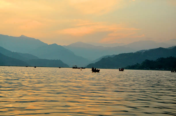 Fewa Lake, Pokhara, captured during a serene sunset, with golden reflections on the water and silhouettes of boats scattered across the tranquil expanse, surrounded by layers of distant hills.