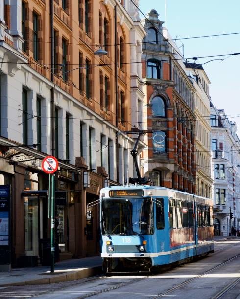 A blue tram travels through a historic street in Oslo, Norway, surrounded by classic European architecture under a clear sky.