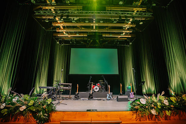 istock Stage setup for a live performance with musical instruments, microphones, a drum set, green lighting, and floral decorations in an auditorium. 2192354955