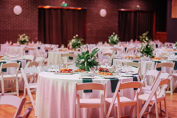 istock Elegant event hall with round tables set for dining, featuring white tablecloths, floral centerpieces, and arranged tableware on wooden flooring. 2192354959