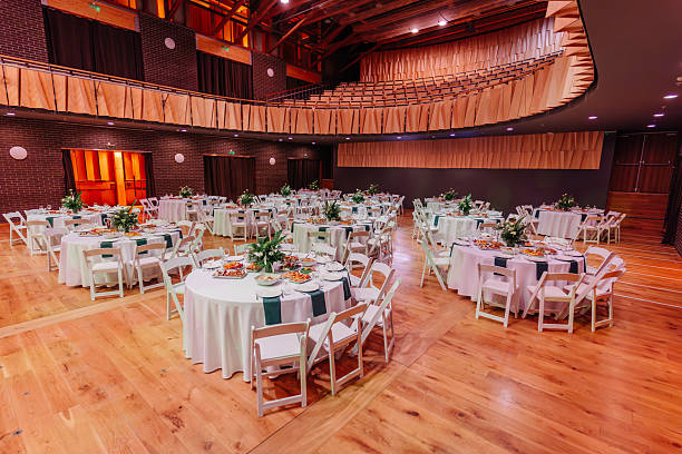 Banquet hall with round tables arranged for an event, decorated with white tablecloths, green napkins, and floral centerpieces on a wooden floor.