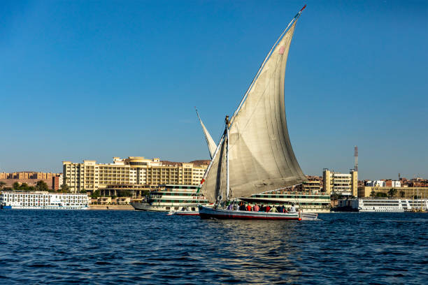 Aswan High Dam, Egypt: A felucca glides on the Nile River in spring, with sandy hills and boats along the shore under a blue sky.