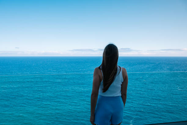 A woman stands on a balcony overlooking the ocean. The sky is clear and blue, and the water is calm. The woman is lost in thought, taking in the beauty of the scene