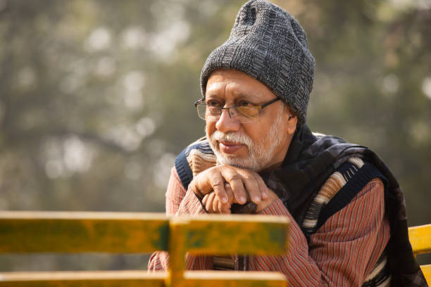 Side view of sad contemplative senior man in warm clothes looking down and sitting on bench in park during weekend