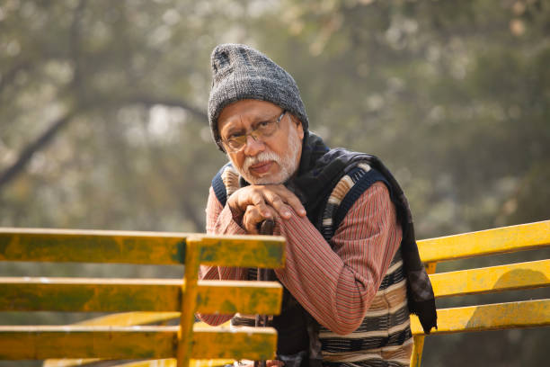 Side view of sad contemplative senior man in warm clothes looking down and sitting on bench in park during weekend