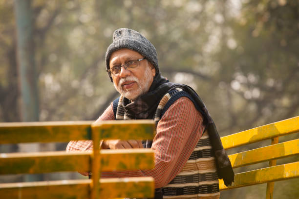 Senior man in warm clothes looking at camera and sitting on bench in park during weekend