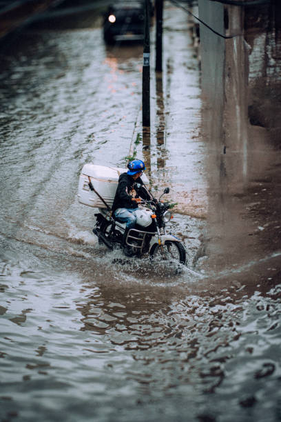 A motorcyclist with a large delivery box braves a heavily flooded street in Cancun, Mexico, following tropical rainfall. The water splashes around the wheels, highlighting the challenges of transportation during extreme weather. This image reflects resilience and the day-to-day struggles in tropical urban areas.