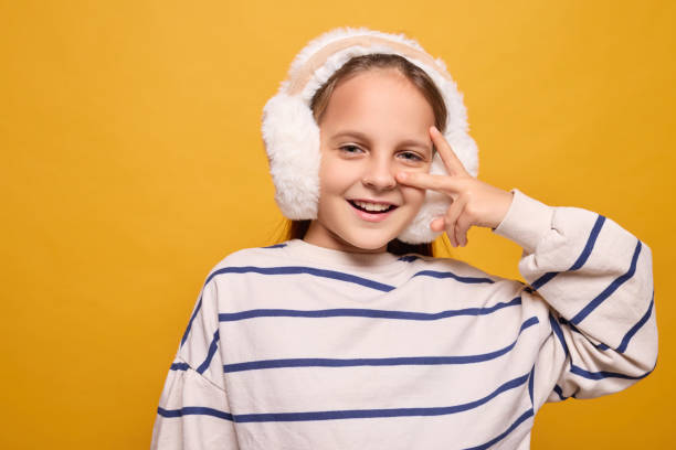 istock A girl enjoys winter, wearing white earmuffs and a striped sweater while playfully posing with her fingers. She exudes joy against a vibrant yellow wall 2192410849