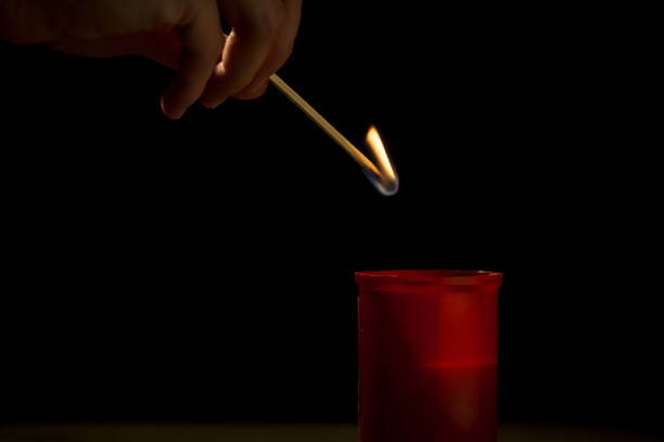 Male hand, using a safety match, lighting a red cemetery candle, on a black background, close up shot. All saints day concept.
