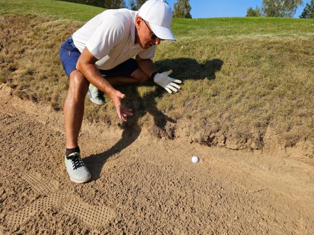 A male golfer swings from a sand bunker, striking the ball as sand lifts into the air
