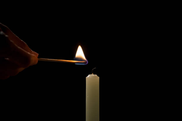 istock Male hand lighting a white thin taper candle with a match on a black background 2192414765