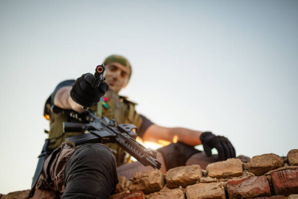 Close-up photo of a military individual aiming a firearm downward, wearing tactical gear on a brick rubble backdrop under a clear sky.