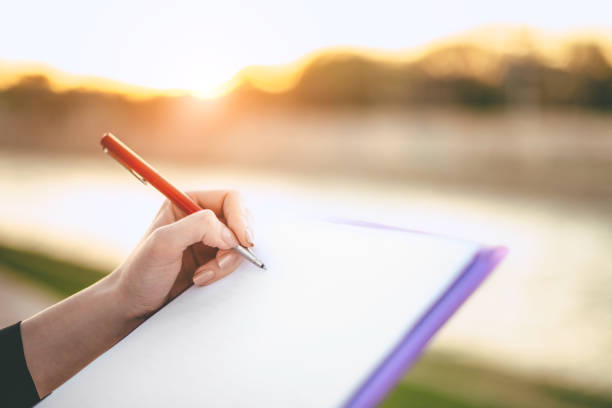A close-up of a hand writing with a pen on a notebook during sunset, showcasing outdoor reflection and creativity, highlighting writing and documentation in a tranquil setting