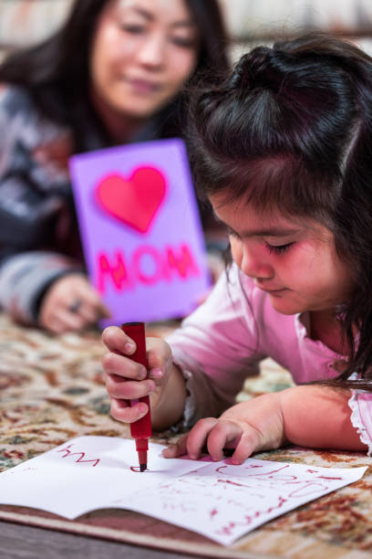 A little girl is drawing a card for her mom. She is using a red marker to write the words \