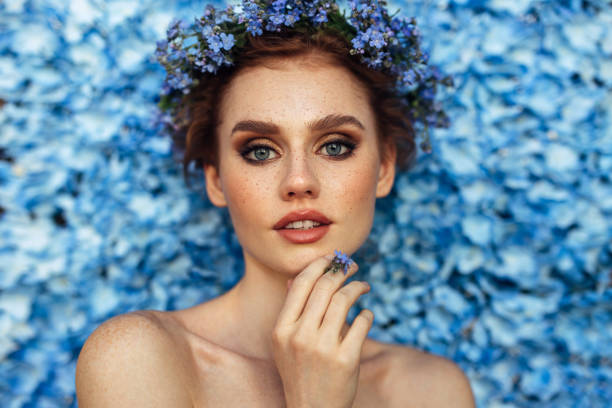 A striking woman with piercing blue eyes and delicate freckles poses gracefully against a sea of vivid blue flowers. A floral crown of the same hue sits atop her soft waves, accentuating her serene beauty. Her hand lightly touches her chin, adorned with a matching floral ring, adding to the harmony of the scene. The image radiates elegance, blending natural beauty with a touch of romance and artistry.