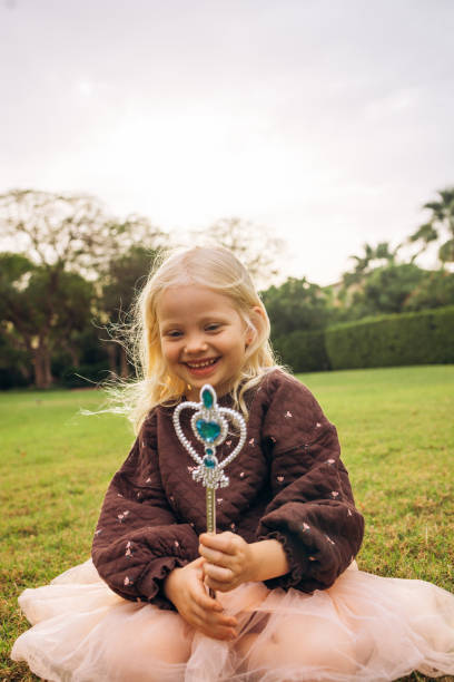 An enchanting photograph of a young blonde girl sitting on a grassy field, holding a wand in her outstretched hand as if casting a spell. The child is dressed in a brown quilted sweater adorned with small patterns and a fluffy peach tutu that adds a touch of whimsy. Her focused expression and the way she holds the wand bring out her imaginative spirit. The background features lush green trees and a hint of a building, creating a serene outdoor setting. This image beautifully captures the essence of childhood fantasy and the simple joy of make-believe.