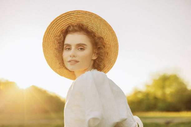 A young woman with curly hair stands gracefully in a sunlit field, wearing a flowing white dress and a straw hat. The golden hour light casts a warm, ethereal glow, highlighting her serene expression and the natural beauty of the scene. Her relaxed pose and the surrounding landscape evoke a sense of peace and timeless elegance.