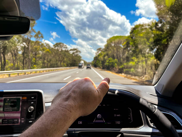istock A wide-angle view through a car’s windscreen reveals a highway through a forested area, framed by the interior, steering wheel, and driver’s hands under a clear blue sky with fluffy clouds 2192440031