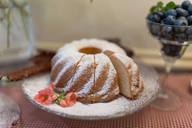 A delectable ring-shaped cake, dusted with powdered sugar, garnished with delicate pink flowers, presented on vintage glassware, with blueberries nearby, offers a glimpse into the culinary charm of a boutique hotel.