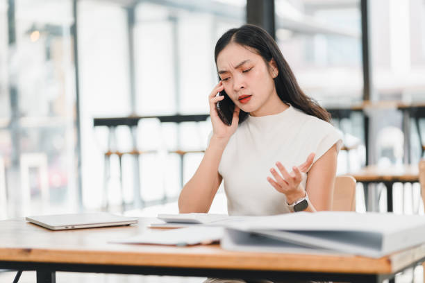 Frustrated woman talking on phone in modern office, expressing concern and confusion while managing paperwork. Her focused demeanor highlights stress of multitasking