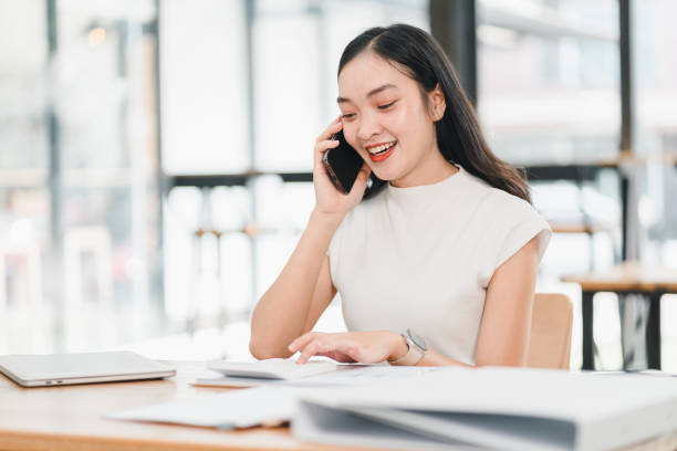 istock Young woman talking on phone while working at desk, smiling and engaged. She is surrounded by papers and laptop, showcasing productive work environment 2192451748