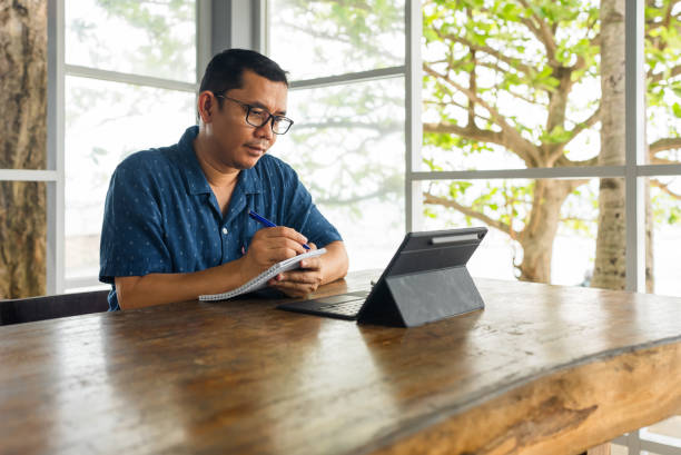 Asian man writing on a book and using laptop stock photo