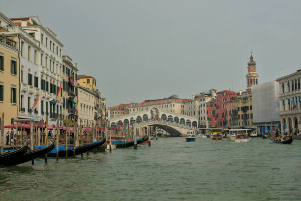 Venice, Italy - October 15, 2024: Venice's Grand Canal sparkles under calm skies. The iconic Rialto Bridge connects both sides of the waterway. Gondolas and boats glide past colorful historic facades.