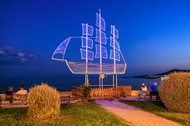 A large illuminated boat is on display in a park by the water. A couple is sitting on a bench nearby. August 10, 2024 Kavala Greece
