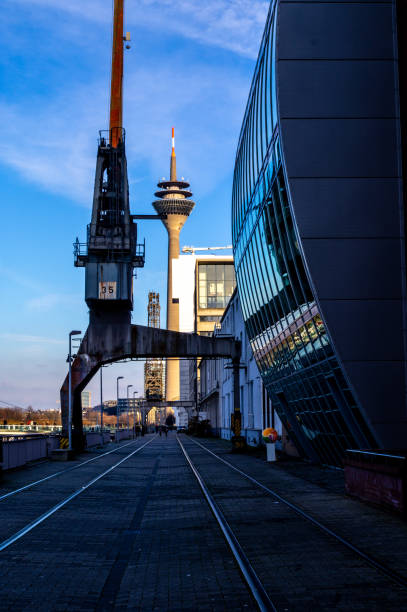 A striking perspective of Düsseldorf's MedienHafen, featuring an industrial crane, tram tracks, and the iconic Rhine Tower (Rheinturm) in the background. The sleek modern architecture and reflections on the glass facade contrast with the historical industrial elements, creating a unique urban atmosphere.
