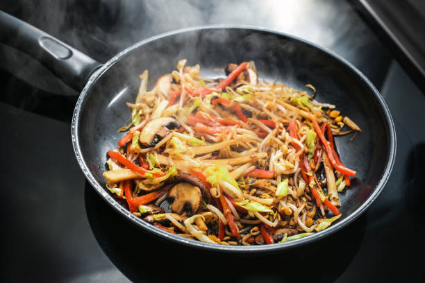 Steaming vegetables stir fried in a black pan, such as mushrooms, red bell peppers, soybean sprouts and cabbage for cooking an Asian meal, selected focus