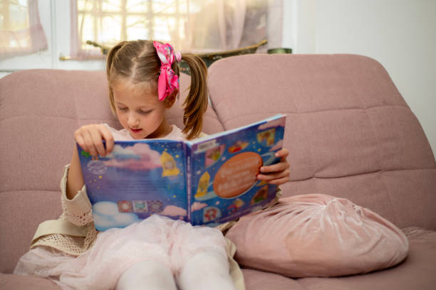 A young girl dressed in light pink clothing sits comfortably on a plush pink couch, absorbed in reading a colorful and engaging children's book. The scene conveys a sense of curiosity and learning in a cozy and serene environment, highlighting the importance of reading and childhood exploration.