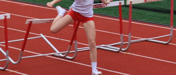 Side view of athlete young woman jumping over hurdles during practice in sports hall.