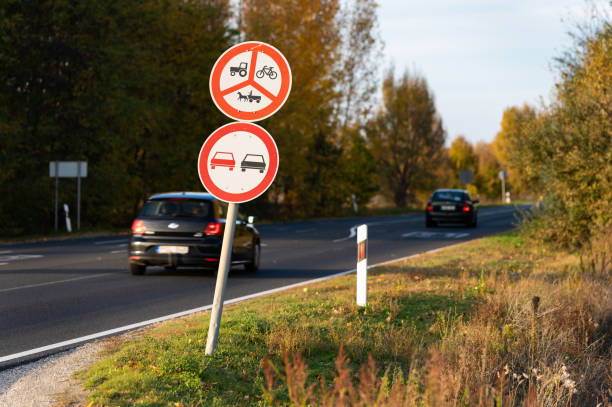 A pair of traffic restriction signs on a rural road prohibiting tractors, bicycles, horse-drawn vehicles, and overtaking. Cars are visible traveling on the curving road surrounded by autumn trees and greenery.