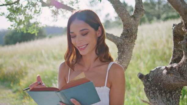 Rural woman writing notes in notebook with pencil portrait. Relaxed girl painting sketch of nature looking camera at vacation. Smiling female model posing in field. Young lady taking break outdoor