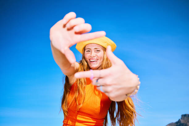 Woman wearing orange sleeveless top looking at camera through square shape, gesturing with hands, pretending, view through, fun