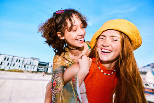 Young woman wearing yellow sunhat giving female friend piggyback stock photo
