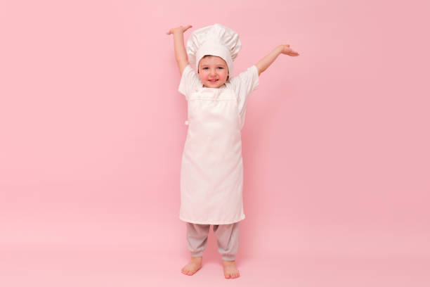 istock Child dressed as a chef in white uniform with arms raised. Studio portrait on pink background. Cooking and culinary concept for poster and card design. Full-length photography. 2192499292