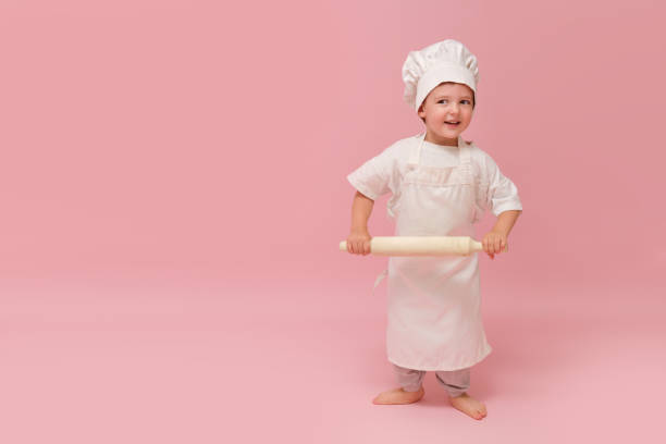 istock A child in a chef's uniform, complete with a hat and apron, is seen holding a rolling pin in a pink-themed studio portrait. Kid aged 3 years (three year old boy) 2192499293